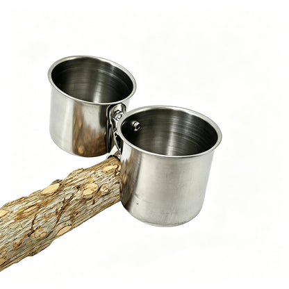 Two stainless steel feeding cups with attached metal clips, shown resting on a textured log against a plain white background.